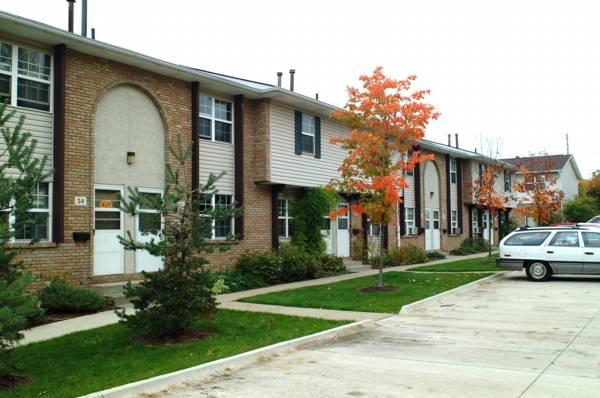 Row of two-story brick townhouses with small front lawns, a few trees with autumn leaves, and a white car parked in a concrete lot.