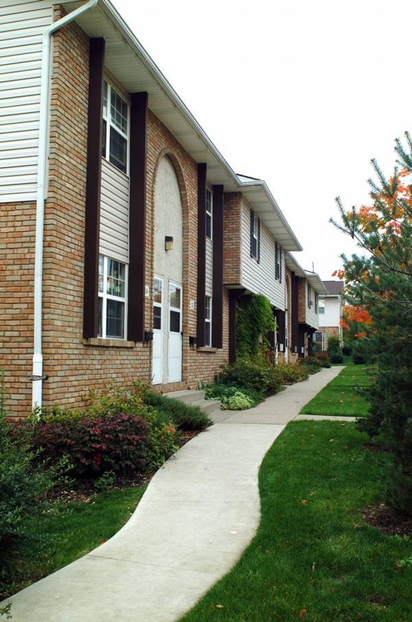 A sidewalk curves past a row of two-story brick apartment buildings with white trim, brown columns, and landscaped bushes and grass.