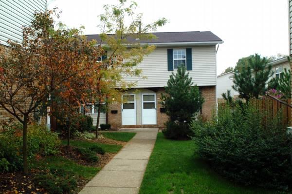 A two-story brick and siding duplex with two front doors, surrounded by green grass, trees, and shrubs along a concrete walkway.