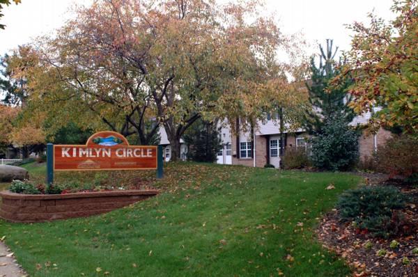 A wooden sign reading "Kimlyn Circle" stands on a grassy lawn with trees and a brick apartment building in the background.