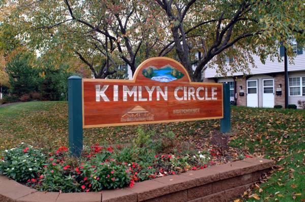 A wooden sign reading "Kimlyn Circle" stands amid flowers and grass, with trees and residential buildings in the background.