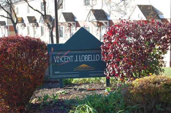 A green sign reading "Vincent J. Lobello" stands among bushes in front of a residential building.