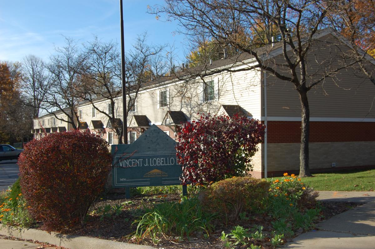A row of tan and brown townhouses with a green sign in front reading "Vincent J. Lobello." Leafless trees and shrubs are present, with some autumn foliage visible.
