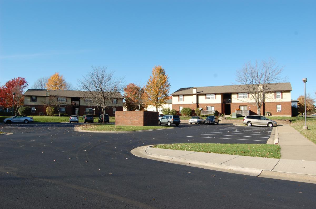 An apartment complex with two-story brick buildings, a mostly empty parking lot, several parked cars, and trees with autumn foliage under a clear blue sky.