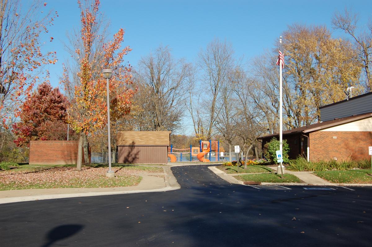 A paved parking lot leads to a playground with orange slides, surrounded by trees with autumn leaves and a building to the right.