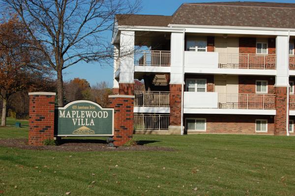 A green sign for Maplewood Villa stands in front of a three-story red brick apartment building on a sunny day.
