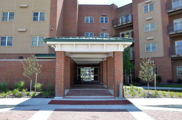 Brick apartment building with covered entrance, small landscaped area, and windows; balconies visible on the right side.