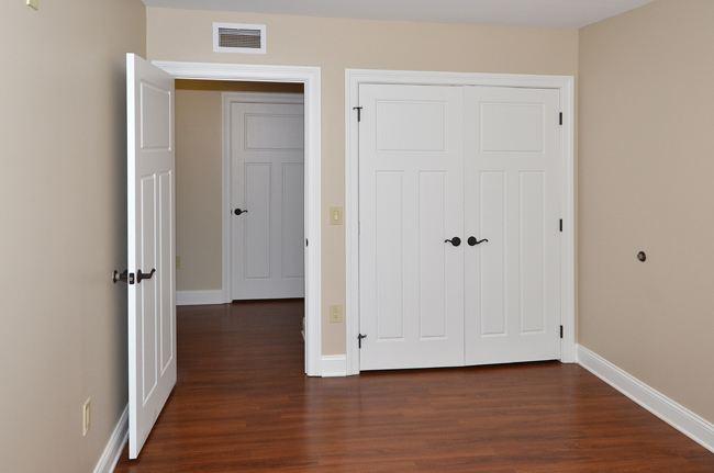 Empty room with beige walls, wood flooring, an open door leading to a hallway, and closed double closet doors.