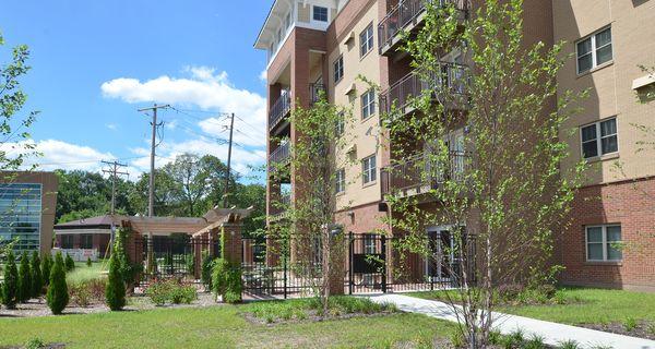Four-story apartment building with balconies, a landscaped yard, young trees, and a paved walkway on a sunny day.