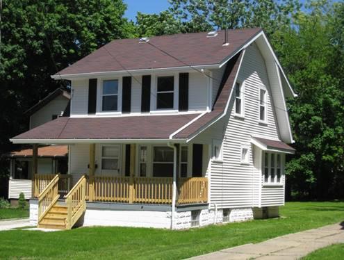 Two-story white house with a red-brown roof, black shutters, and a covered front porch with wooden railings, set on a grassy lot with trees in the background.