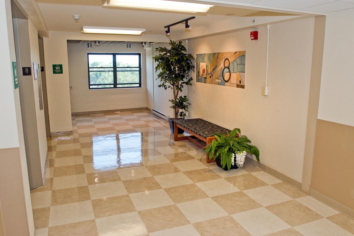 A hallway with checkered tile flooring, a bench with cushions, two potted plants, a window at the end, and wall art above the bench.