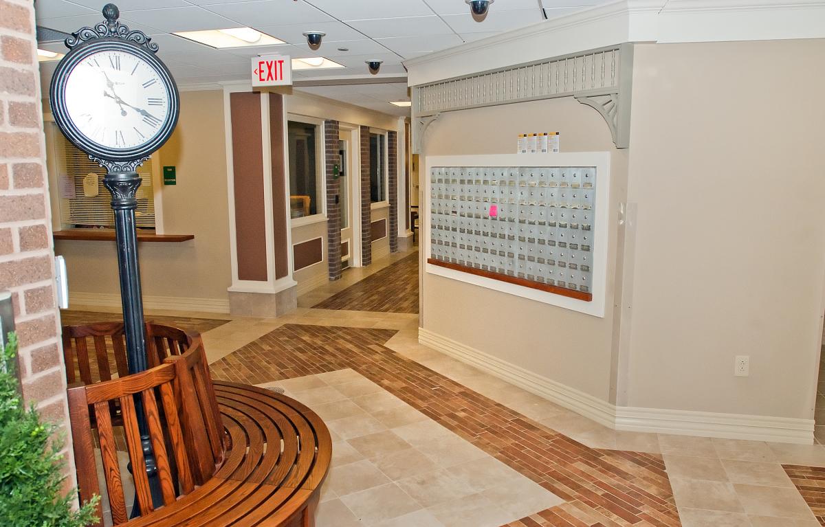 Hallway with a wall of mailboxes, a wooden bench with a black clock, tiled floors, and an exit sign on the ceiling.
