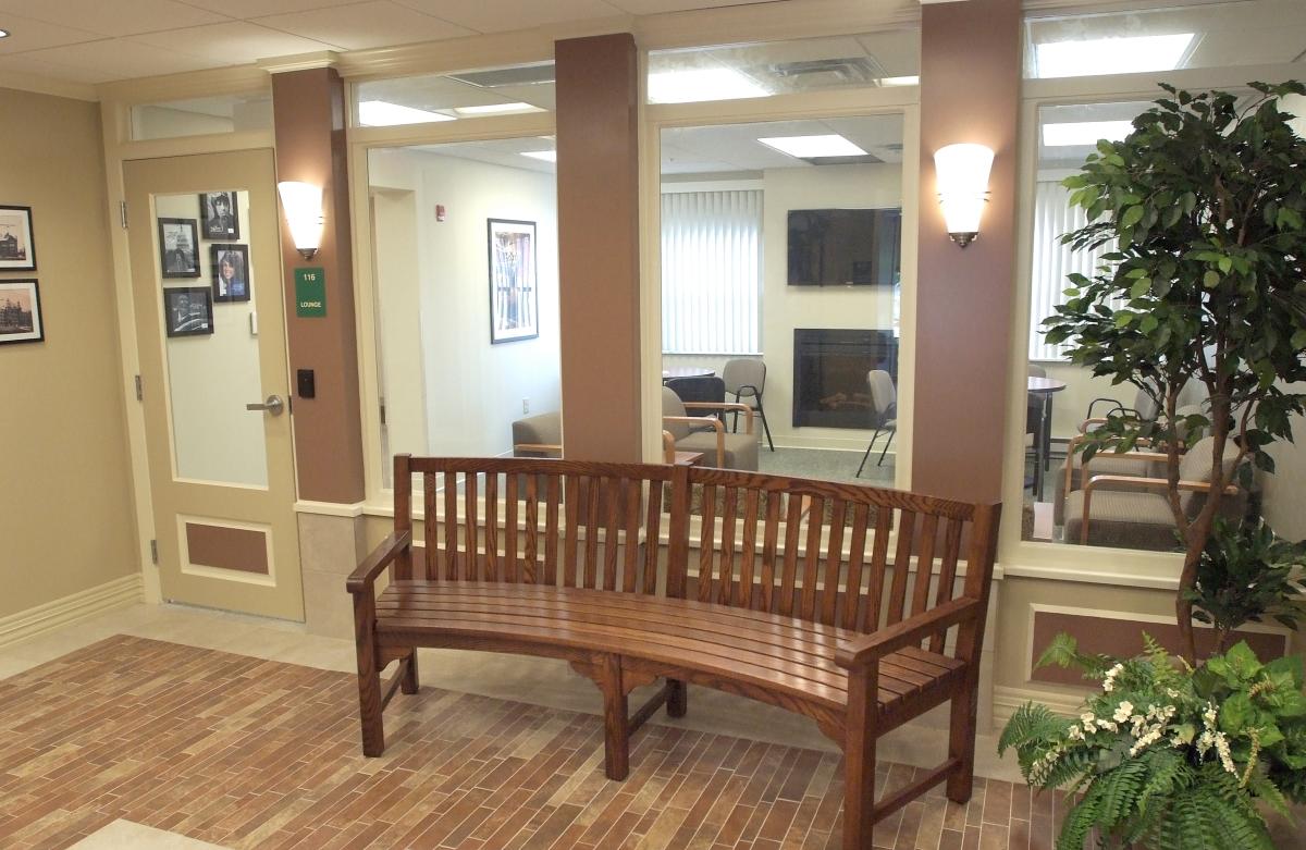 A curved wooden bench sits in a hallway with brick flooring, wall-mounted lights, framed photos, and large glass windows looking into an office or meeting room.