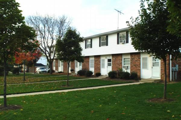 A two-story brick and white siding apartment building with multiple entrances, surrounded by grass, trees, and a sidewalk.