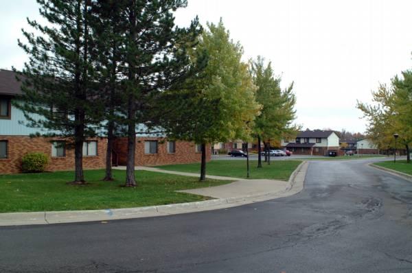 A paved road curves past apartment buildings with brick exteriors, surrounded by green grass, trees, and overcast sky.