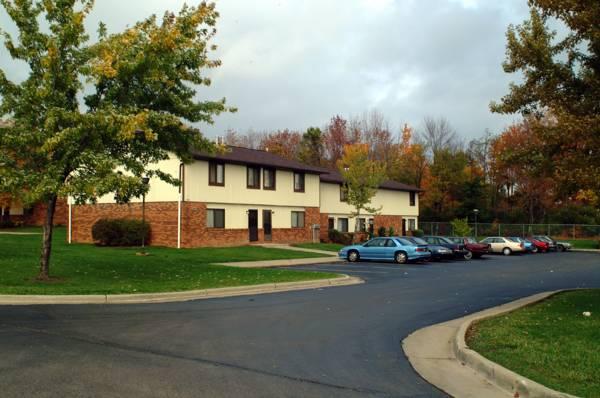 Two-story apartment building with a brick and beige exterior, surrounded by trees, a grass lawn, and several parked cars in a lot on a cloudy day.