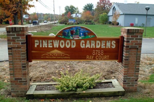 Wooden sign reading "Pinewood Gardens" with address "8788 Ray Court," supported by brick pillars, with some small plants in front and a street in the background.