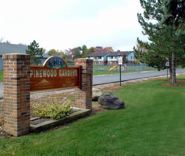 A brick sign for Pinewood Gardens stands on a lawn near a playground, with houses and trees visible in the background under a cloudy sky.