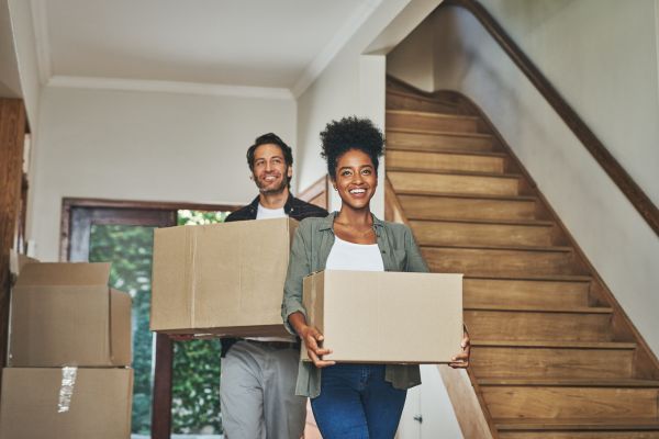 Two people smile while carrying cardboard boxes inside a house with a wooden staircase and more boxes visible in the background.