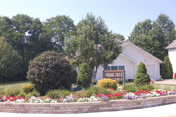 Single-story house with a small front yard, flower bed, and a sign reading "POULIAC CIRCLE." Large trees and bushes surround the area under a clear sky.