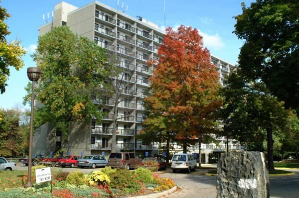 A multi-story apartment building with balconies is surrounded by trees, parked cars, and a landscaped garden under a blue sky.