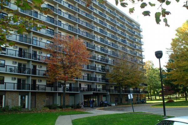A multi-story apartment building with balconies, surrounded by trees with autumn foliage and a sidewalk in the foreground.