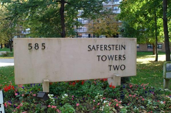 A beige sign reads "585. Saferstein Towers Two," surrounded by flowers, with trees and an apartment building in the background.