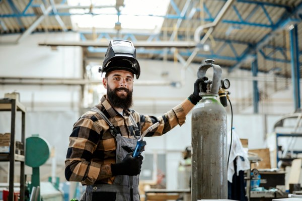 A person wearing a welding helmet and gloves stands in a workshop, holding a tool and resting a hand on a large gas cylinder.