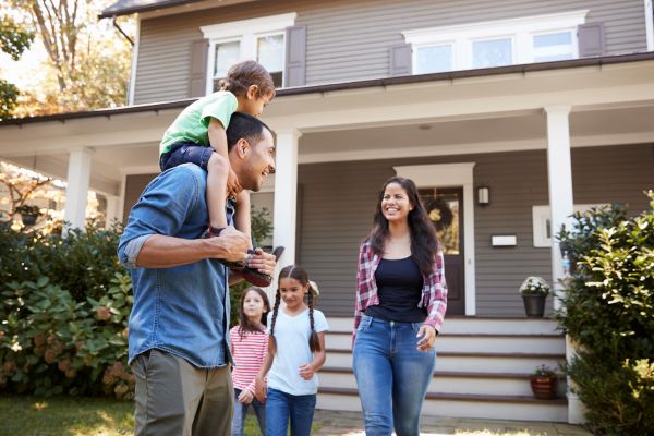 A man carries a child on his shoulders while walking with two other children and a woman in front of a house with a porch.