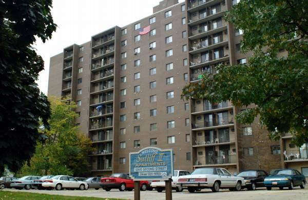 A multi-story apartment building with balconies, parked cars in front, and a sign reading "Shiloh Apartments, 8000 Saginaw Street." Trees frame the foreground.