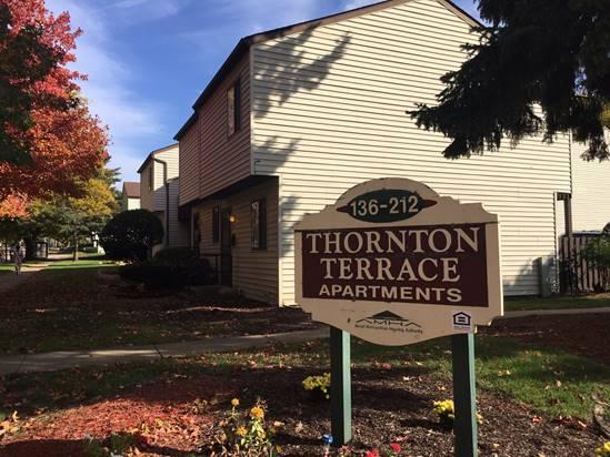 A brown and white sign reads “Thornton Terrace Apartments 136-212” in front of a two-story apartment building with trees and landscaping.
