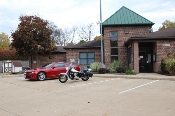 A red car and a motorcycle are parked in front of a brick building with a green metal roof. The parking lot is mostly empty.