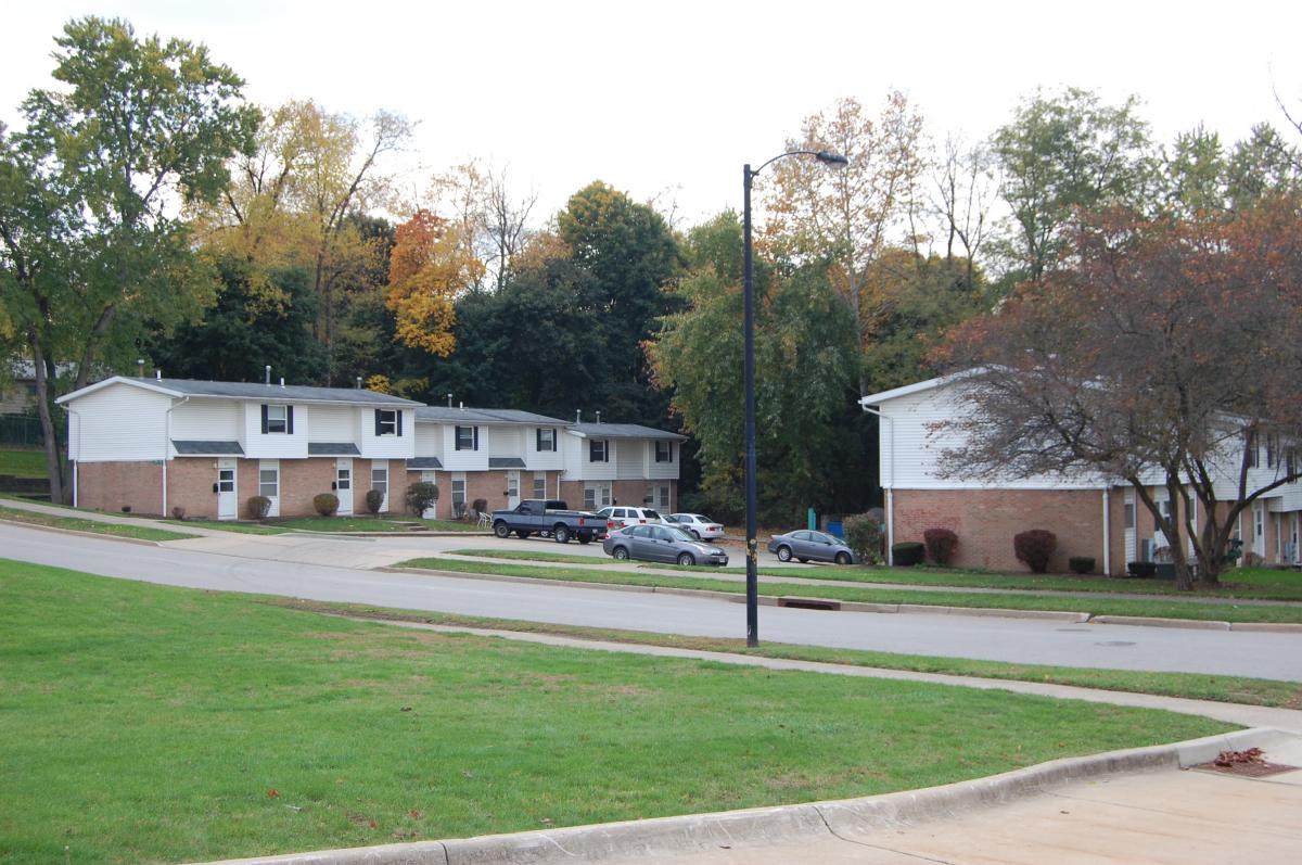 Two brick apartment buildings with parking spaces and cars in front, surrounded by grass, trees, and a street curving through the residential area.