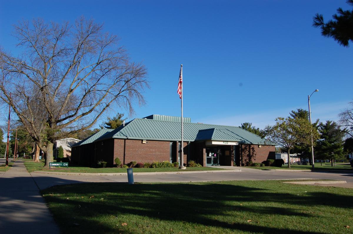 A single-story brick building with a green metal roof, an American flag on a pole in front, surrounded by grass, trees, and a sidewalk under a clear blue sky.