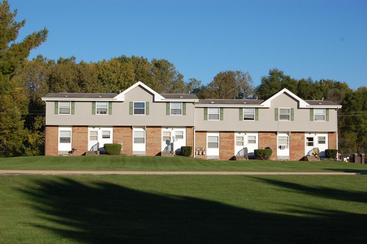 A row of two-story townhouses with beige siding and brick exteriors sits on a well-kept lawn with trees in the background under a clear blue sky.