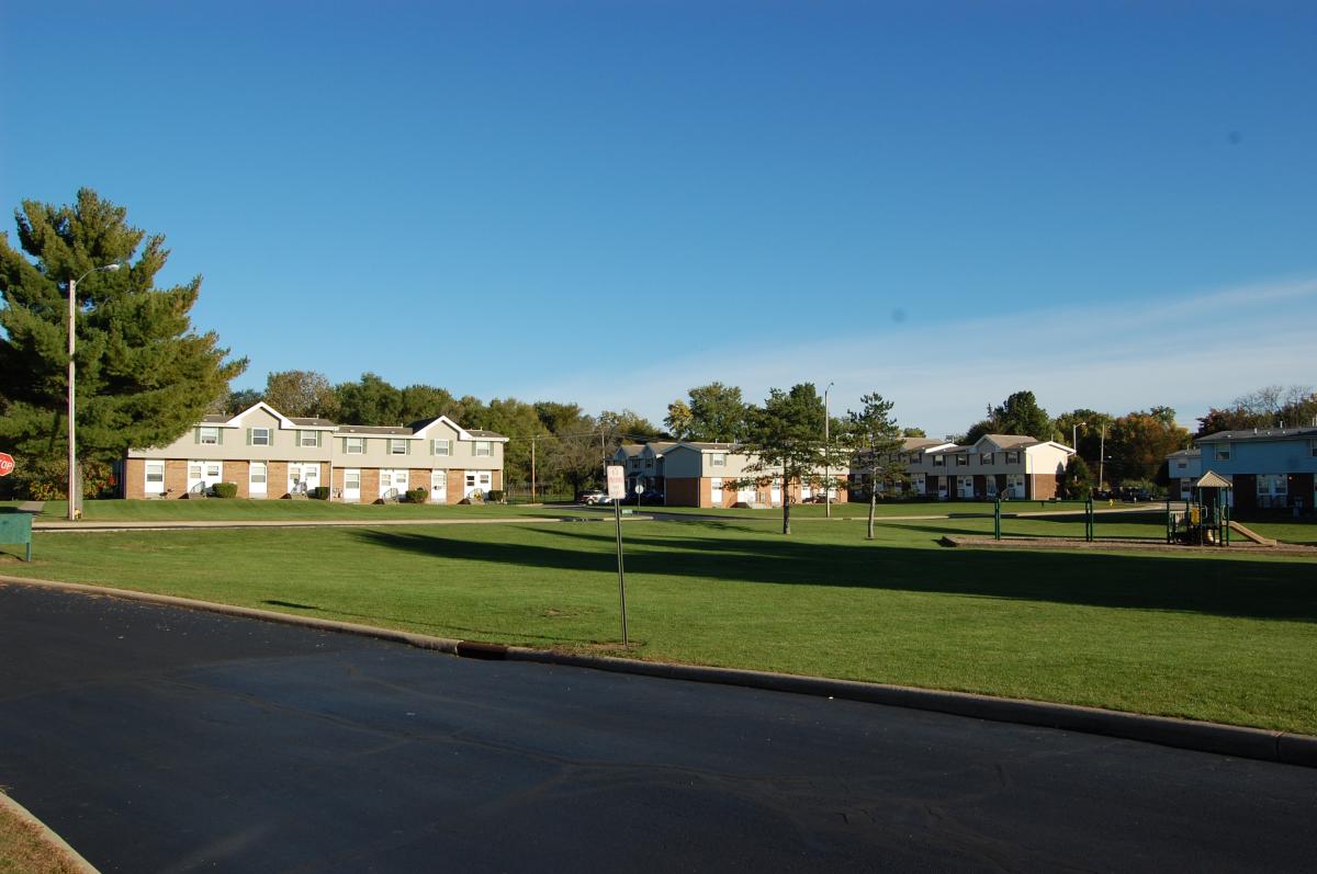 Apartment buildings border a large grassy lawn with a few trees and a playground, under a clear blue sky.