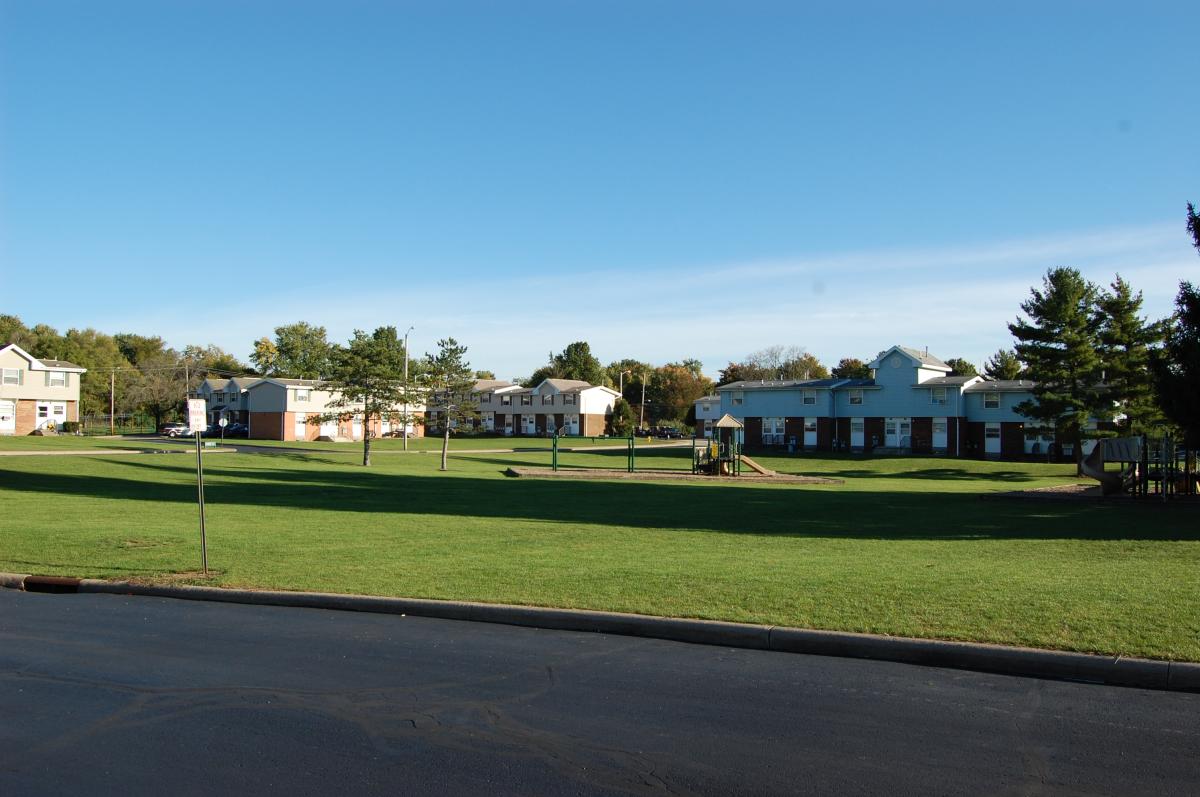 Open grassy area with a playground, bordered by residential buildings and trees under a clear blue sky.