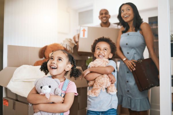 Two children holding stuffed animals stand in front of moving boxes, with two adults smiling in the background, inside a home.