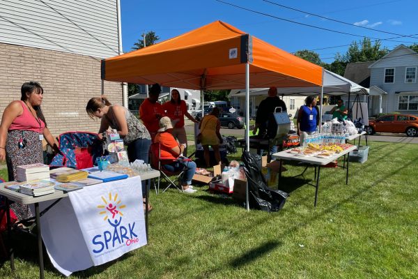 People gather under orange tents and at tables with food, drinks, and books at an outdoor community event on a sunny day.