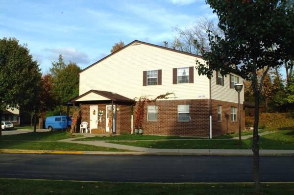 Two-story brick and siding house with a front porch, white chairs, and brown shutters. A blue van is parked on the left, and trees with autumn foliage surround the property.