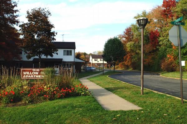 Entrance to Willow Run Apartments with a sign, sidewalk, streetlamp, and autumn trees. A road curves past residential buildings under a partly cloudy sky.